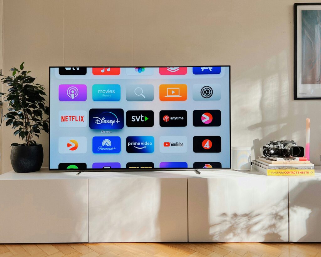 Modern living room with a flat-screen smart TV on a white cabinet displaying streaming app icons including Netflix, Disney+, Prime Video, YouTube, and more. A potted plant sits on the left, and a stack of books with a vintage camera and candle is on the right illustrating IPTV Nordic streaming experience.